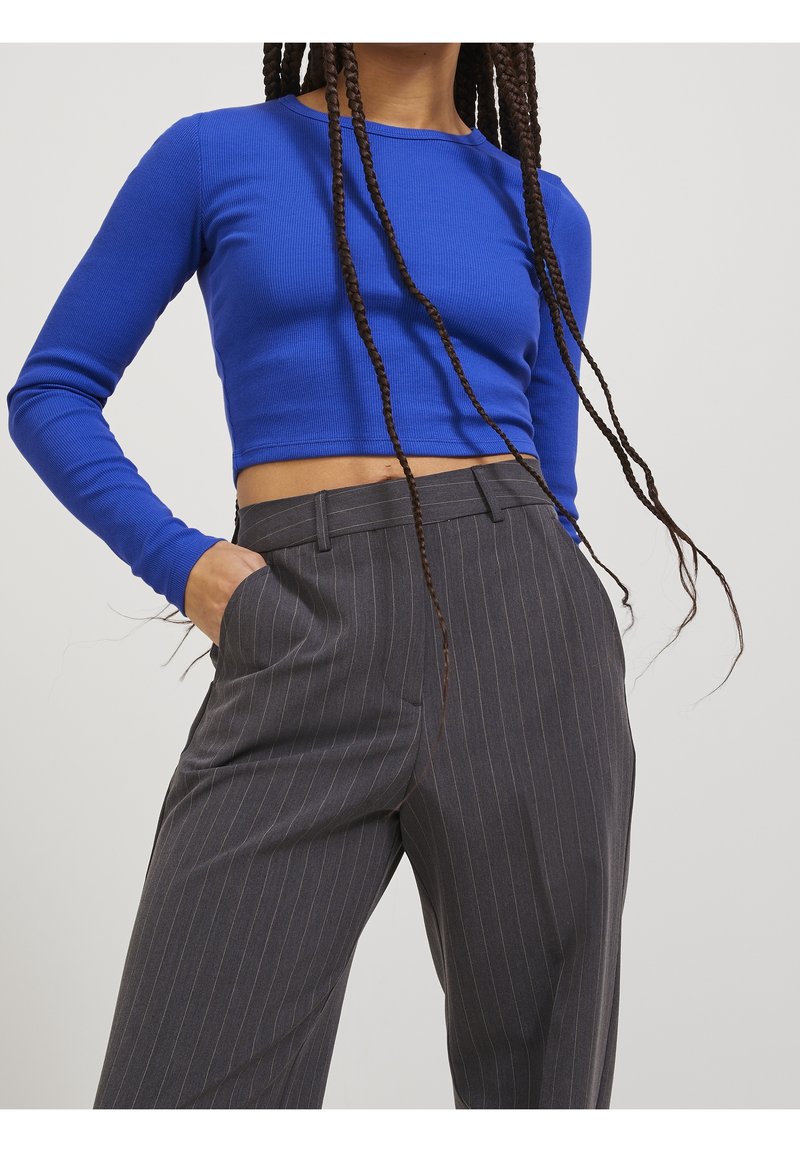 Person wearing a blue long-sleeve crop top and high-waisted gray pinstripe trousers with hand in pocket against a plain background.
