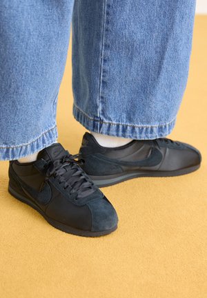 Juodi Nike sportbačiai, pagaminti iš zomšos ir audinių, su apvaliu pirštu, raišteliais ir tekstūruota guma, derinami su mėlynomis džinsomis.