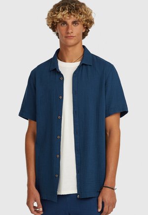 Młody mężczyzna o kręconych blond włosach, ubrany w niebieską koszulę na krótki rękaw z guzikami na białej koszulce, stojący na jednolitym tle.