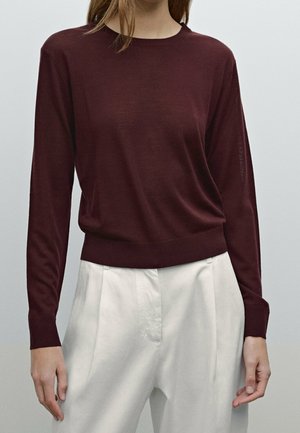 Persona che indossa un maglione aderente a maniche lunghe color bordeaux infilato in pantaloni bianchi a vita alta con pieghe, su uno sfondo semplice.