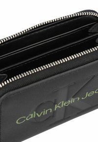 Portafoglio in pelle nera con zip argentata, dotato di molteplici slot per carte e logo verde "Calvin Klein Jeans" in rilievo sul davanti.