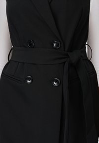 Μαύρο διπλόπατο γιλέκο με τέσσερα κουμπιά και δεμένη ζώνη στη μέση πάνω από λευκό πουκάμισο με μακριά μανίκια, εμφανίζοντας λεπτομέρειες στη μέση.