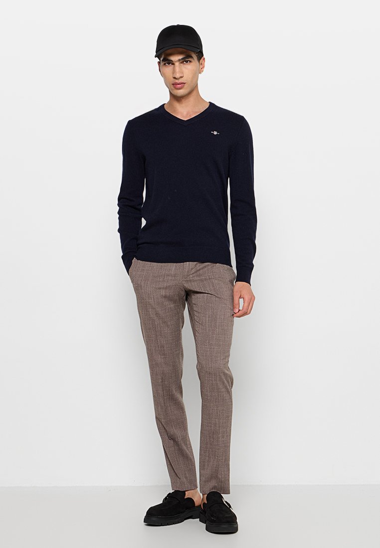 Navy V-Ausschnitt Pullover, gerippte Bündchen, kombiniert mit hellbraunen karierten Hosen. Eine schwarze Mütze und Schuhe vervollständigen den Look.