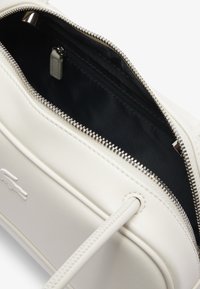 Borsa in pelle bianca con zip argento aperta che mostra l'interno in tessuto scuro e un logo esterno in rilievo a forma di coccodrillo.