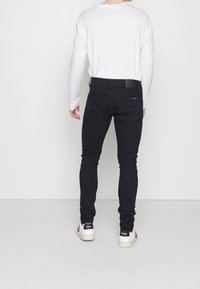 Černé džíny s úzkým střihem a hladkou texturou, s páskem zdobeným koženou nášivkou a dvěma zadními kapsami. Nošeny s bílou košilí s dlouhým rukávem.