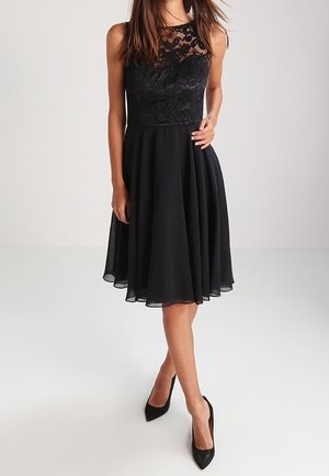 Femme portant une robe noire sans manches avec un corsage en dentelle et une jupe évasée, associée à des talons noirs pointus, debout avec une main sur la taille.