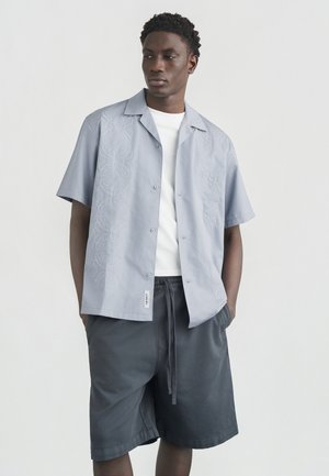 Ung mann iført lyseblå kortermet skjorte med hvit brodering over hvit T-skjorte og løse mørkegrå shorts, hendene i lommene.