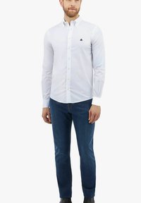 Witte button-up shirt met lange mouwen, voorzien van een klein zwart logo, gecombineerd met blauwe jeans en zwarte laarzen. Effen textuur, kraagdetail zichtbaar.