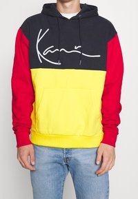 Felpa con cappuccio dal design a blocchi di colore: parte superiore nera, maniche rosse e corpo giallo. Tasca a marsupio davanti e logo in scrittura bianca.