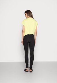 Leggings pretos combinados com uma blusa amarela clara de manga curta e gola alta. A modelo usa sandálias pretas com tiras. Design e ajuste simples.