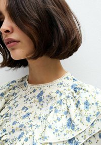 Blusa bianca a fiori con fiori blu, maniche a volant e un delicato scollo in pizzo. Tessuto leggero e arioso con dettagli testurizzati.