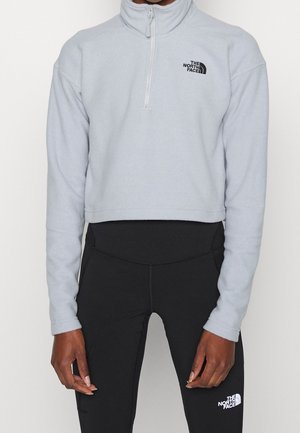 Person iført lys grå kort lynlås-fleece med sort logo og sorte højttaljede leggings med hvid logo, stående mod en ensfarvet baggrund.