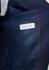 Interior de una chaqueta de traje naval con tela de patrón paisley, costuras de acento rojas, botón negro y una etiqueta que dice "PIER•ONE."