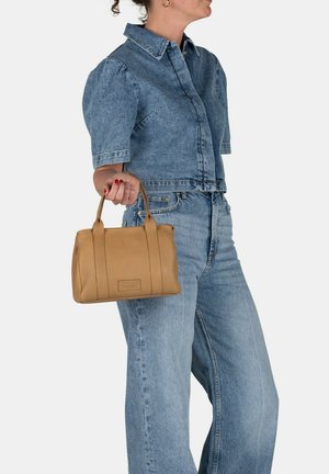 Mujer vestida con un conjunto de mezclilla azul que sostiene un pequeño bolso de cuero color tostado con asas superiores frente a un fondo liso.