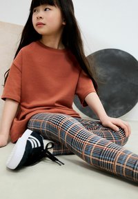 Braunes Baumwoll-T-Shirt mit kurzen Ärmeln, kombiniert mit gemusterten Leggings in Schwarz, Blau und Orange mit kariertem Muster, ergänzt durch schwarze Sneakers.