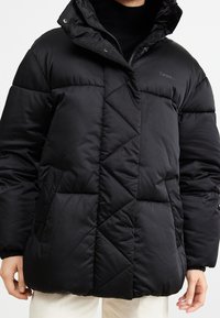 Svart puffjacka med huva, med en quiltad design, blixtlås och två sidofickor. Tillverkad av ett mjukt, glänsande material.