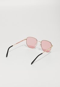 Gafas de sol de metal con lentes rosas, forma hexagonal, delgado marco dorado, almohadillas de nariz transparentes y puntas negras en las varillas.