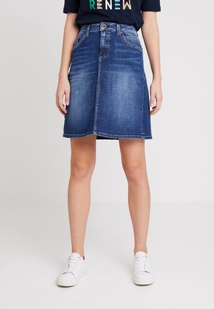 Kobieta ubrana w niebieską dżinsową spódnicę w kształcie litery A, granatowy T-shirt z wielokolorowym napisem "RENEW" i białe trampki, stoi na jednolitym tle.