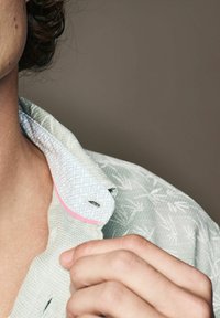 Lys grøn skjorte med mønster i struktureret stof, en blå mønstret krave og pink detaljer. Viser en del af en hånd, der griber fat om kraven.