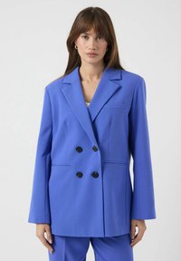 Dobbelspent blazer i lyst blått stoff. Har store svarte knapper, hakkelapper og frontlommer med en skreddersydd passform.