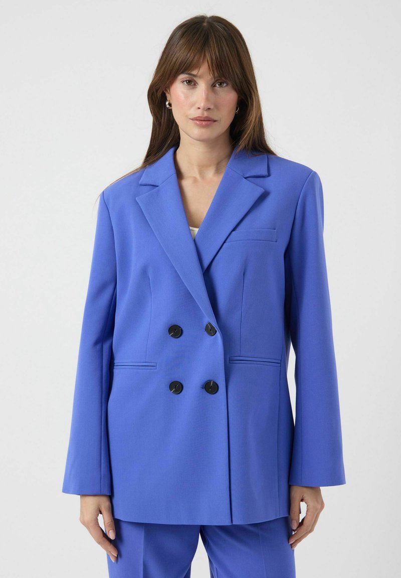 Dobbelspent blazer i lyst blått stoff. Har store svarte knapper, hakkelapper og frontlommer med en skreddersydd passform.