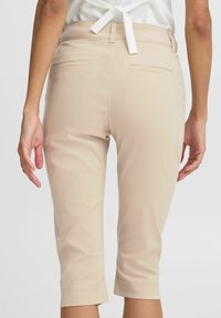 Pantalons capri beiges avec une texture lisse, dotés d'une taille ajustée, de poches arrière et d'ourlets retroussés. La main du modèle repose sur la hanche.