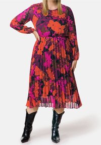 Robe florale violette à manches longues, avec des plis et des motifs orange, rose et noir. Associée à des bottines noires brillantes.