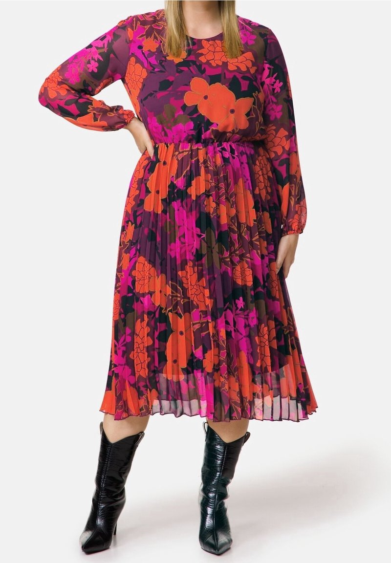 Robe florale violette à manches longues, avec des plis et des motifs orange, rose et noir. Associée à des bottines noires brillantes.