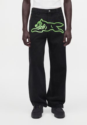 Homme portant un jean noir à jambes larges avec un grand dessin de chien cartoon vert sur la partie avant de la cuisse.