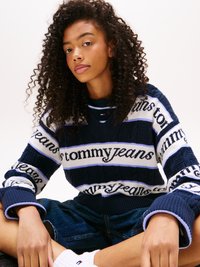 Pletený sveter v námorníckej modrej a bielej farbe s textom "tommy jeans", ktorý má štruktúrovaný dizajn, výstrih do V a rebrované manžety. Viditeľné sú aj deninové šortky.