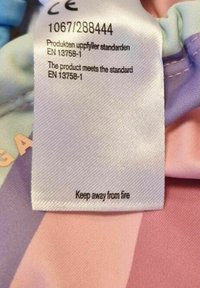 Vaatelappu vaalealla kankaalla. Lappu on valkoista satiinia mustalla tekstillä, joka ilmoittaa CE-merkinnästä ja paloturvallisuushälytyksestä.
