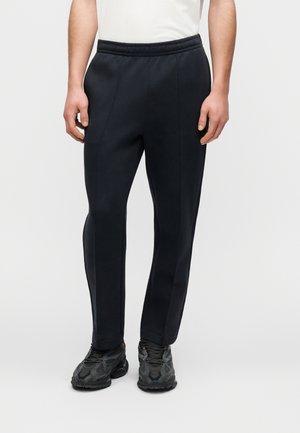 Marineblå joggerbukser med blød tekstur, elastisk talje, tilspidsede ben og subtile sømdetaljer, kombineret med mørke sportssko.