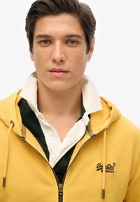 Sudadera amarilla con cremallera y bolsillos frontales y capucha ajustable, superpuesta a una camisa blanca de cuello con un patrón de cuadro verde y negro.