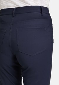 Mörkblå snug-fit byxor med en slät textur. Funktioner inkluderar dubbla bakfickor och en midjeband med bältesöglor.