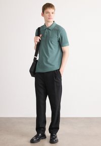 Ung mann står iført en grønn poloskjorte, svarte bukser, svarte loafers, og bærer en svart skulderveske, ser litt til siden.