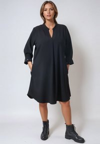 Zwarte A-lijn jurk met een kraag, driekwart mouwen en zijvakken. Glad materiaal met een lichte glans, gecombineerd met zwarte veterschoenen.