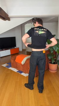 Mężczyzna w czarnej koszulce i ciemnych dżinsach stoi w salonie z drewnianą podłogą, pomarańczową sofą, telewizorem i dużą rośliną doniczkową.