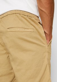 Personne portant un pantalon élastique beige avec des poches latérales et arrière, la main reposant à l'intérieur de la poche latérale.
