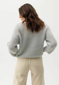 Világosszürke, ribbelt textúrájú kötött pulóver, laza szabással és eső vállakkal, világos bézs nadrággal párosítva.