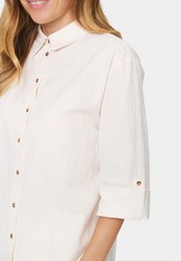 Light beige button-up shirt featuring a collared neckline, rolled sleeves with button accents, and a textured fabric.