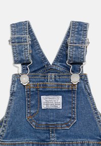 Τζιν μπλε φορμάκι με ρυθμιζόμενους ιμάντες, μεταλλικά κλείστρα, μπροστινή τσέπη και λευκή ετικέτα Levi Strauss & Co. ραμμένη στην τσέπη στο στήθος.