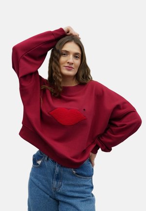 Kobieta o falowanych włosach ubrana w czerwoną bluzę z teksturowanymi ustami oraz niebieskie dżinsy, pozująca z jedną ręką na głowie, a drugą na biodrze.
