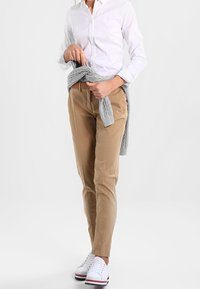 Beige Hose im Slim-Fit, mit glatter Textur und geraden Beinen. Kombiniert mit einem weißen Hemd und einem grau gebundenen Pullover um die Taille.