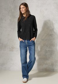 Schwarzes langärmliges Oberteil mit geometrischem Muster, kombiniert mit hellblauen weit geschnittenen Jeans mit Knopfreihe und weißen Turnschuhen.