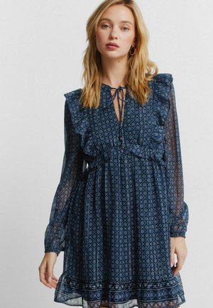 Vaaleahiuksinen nainen, joka käyttää tummansinistä kukkakuosista mekkoa, jossa on röyhelöitä, pitkät läpikuultavat hihat ja kaulanauha, seisoo yksinkertaisen taustan edessä.