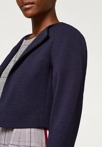 Blazer corto blu navy con texture liscia e colletto a rilievo, abbinato a un vestito grigio a quadri con accenti rossi.