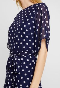 Mörkblå klänning med prickar, transparanta svärmarmer och en figurnära midja, utrustad med ett knytbälte och lätt material.