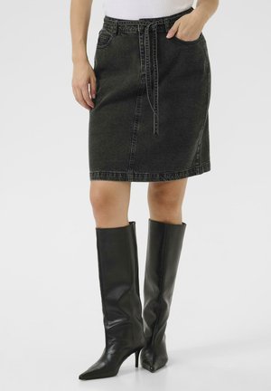 Jupe en denim noir à coupe droite, avec poches avant et une ceinture à nouer à la taille, assortie à des bottes noires en cuir pointues montantes jusqu'aux genoux.