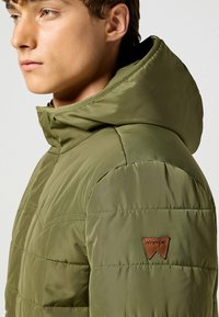 Chaqueta de plumas verde oliva con capucha desmontable, textura acolchada y un parche de cuero marrón con el logo de Wrangler en la manga.