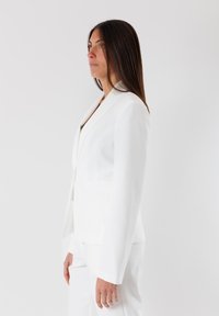 Blazer bianco con una silhouette aderente, spalle strutturate e rever a punta. Tessuto liscio con tasche laterali, abbinato a pantaloni bianchi.
